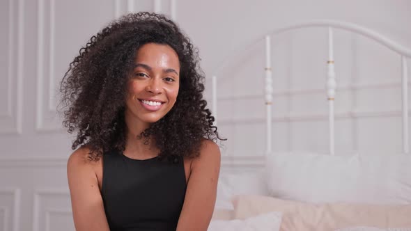 Black Woman Shakes Her Afro Hair with Hands and Looks at Camera Sitting on Bed