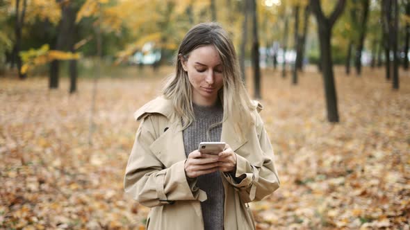 Young Blonde Woman Writing Message on Cell Phone in a Autumn Park