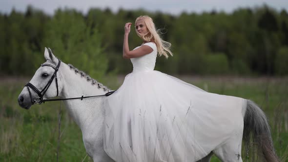 Happy Blonde Bride is Riding Horse Over Meadow at Summer Beautiful Woman on Horseback