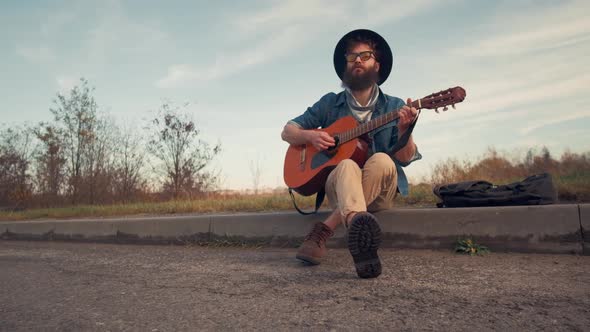 Young Man Playing Guitar and Sitting on Ground in Urban Road