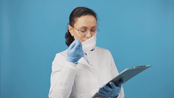 Female Medical Doctor Taking Off Protective Mask and Smiling at Camera Holding Clipboard Blue Studio