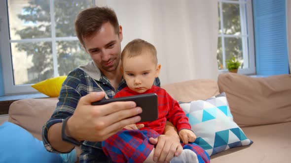 Happy Dad with Little Son Using Smartphone Sitting on Couch