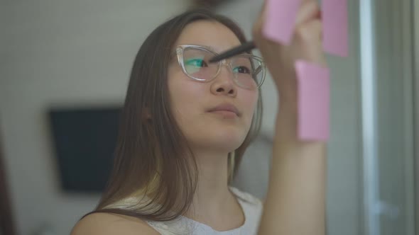 Reflection in Mirror of Confident Slim Young Asian Woman in Eyeglasses Writing with Pen on Sticky