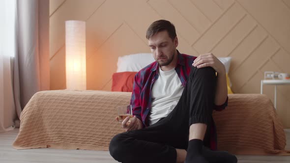 Depressed Man is Sitting on Floor with Glass of Alcohol in Hand with an Absent Look Front View