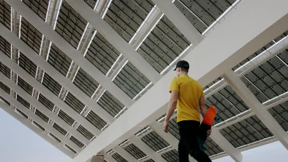 Young Attractive Trendy Man walking with a skateboard under a solar panel on a morning sunny day wit