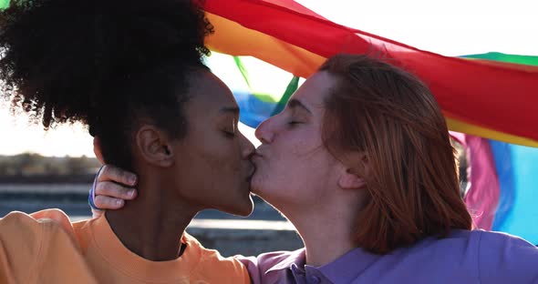 Multiracial lesbian girls having tender moment outdoor wearing LGBT rainbow flag