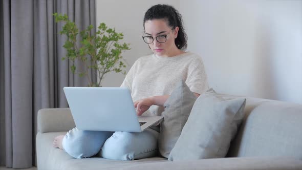 Young Serious Woman Freelancer Working on a Laptop Sitting on a Sofa at Home, Businesswoman Typing