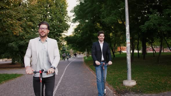 Portraits of Two Handsome Young Businessman Riding an Electric Scooters on the Road in City Park