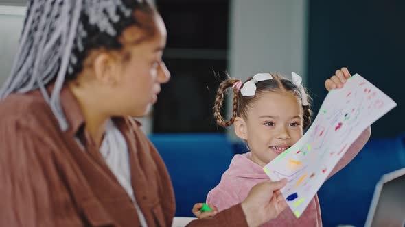 Cute Little African American Girl Showing Colorful Picture to Busy Mother Woman Working on Laptop at