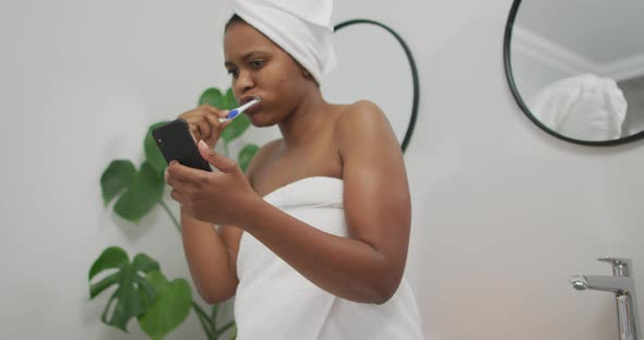 Happy african american woman brushing teeth and using smartphone in bathroom
