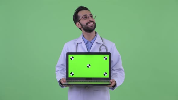 Happy Young Bearded Persian Man Doctor Thinking While Showing Laptop