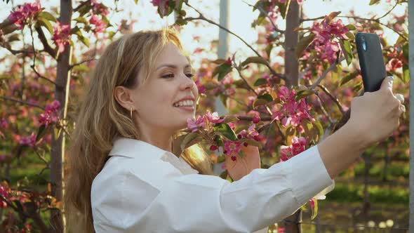 Young Woman in a White Blouse Takes a Selfie Near the Young Trees with Pink Bloom