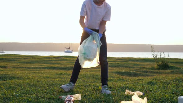 Teenage Girl Collects Rubbish in Gloves on the River Bank at Sunset