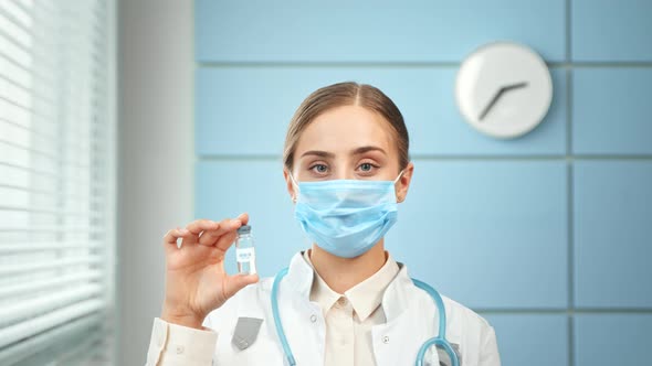 Young woman doctor in blue disposable face mask and white coat holds vial