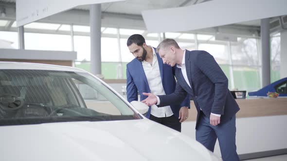 Two Adult Men Discussing New White Car in Dealership. Caucasian Dealer and Middle Eastern Client