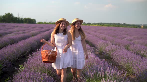 Two Caucasian Women Travelers are Walking in a Lavender Field in Light Summer Dresses and Straw Hats
