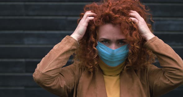 Beautiful Young Red-haired Caucasian Woman Girl in Medical Mask Looking Into the Camera Touch Hair