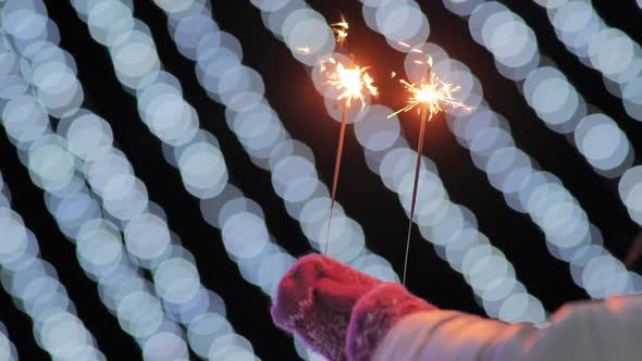 Sparklers in the Hands of a Girl in Mittens Against the Background of the City's Christmas Lights