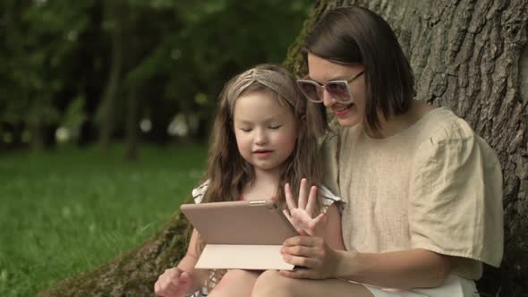 Young Woman with a Little Daughter is Sitting in the Park Under a Large Old Tree and Looking at the