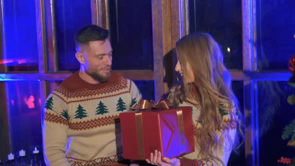 Lovely couple in Christmas sweaters. Handsome man giving a red box with present to beloved girl.