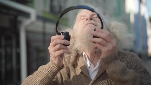 Close-up Portrait of Joyful Bearded Caucasian Man with Long Grey Hair Putting on Headphones and