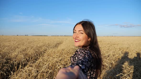 Brunette Woman Leads Across a Field of Ripe Wheat Turning Smiling