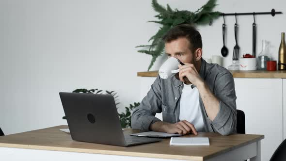 A Young Thoughtful Freelance Man Looks at the Monitor Screen, Drinks Coffee or Tea