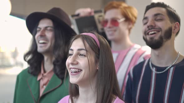 Cheerful Young Slim Woman with Braces Laughing Out Loud Looking at Camera with Blurred Men at