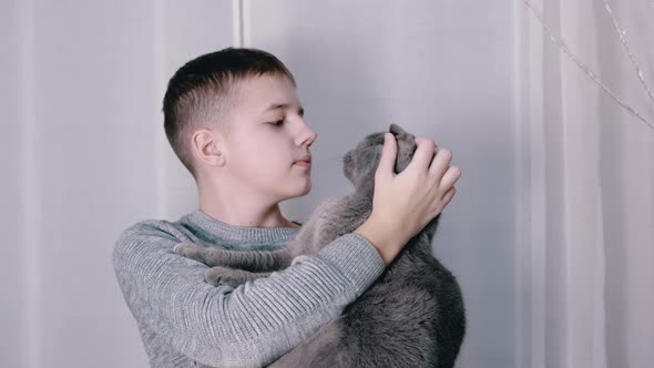 Smiling Boy Hugs Kisses a Fluffy Cat in His Arms in Room