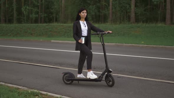 Business Woman Standing with Electric Scooter Wearing Black Suit and Holding Cup of Coffee