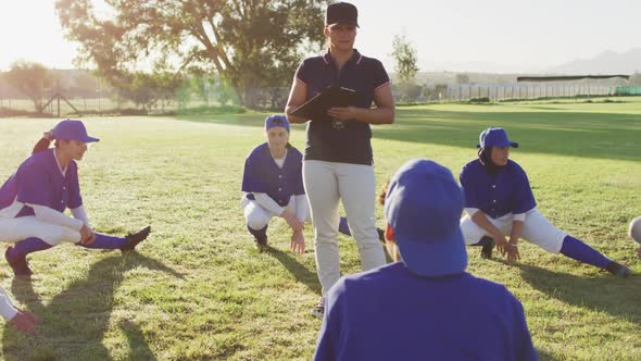 Diverse group of female baseball players with coach, warming up on pitch, squatting, stretching legs