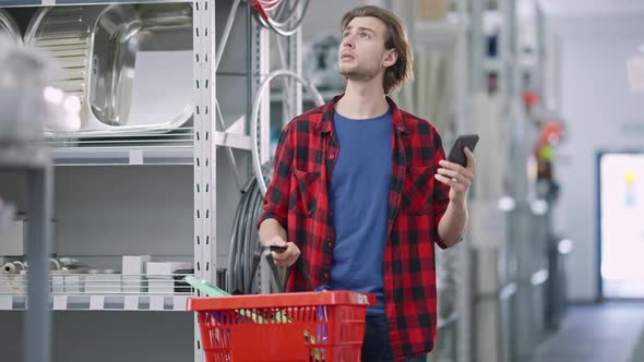 Concentrated Young Man Checking List of Purchases in Smartphone with Goods in Shopping Basket