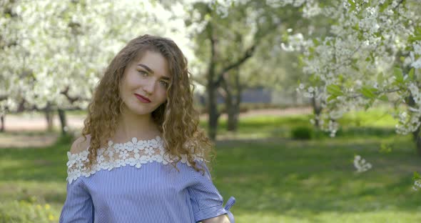 Charming Carefree Girl with Curly Hair Is Standing in Blooming Garden in Spring Day