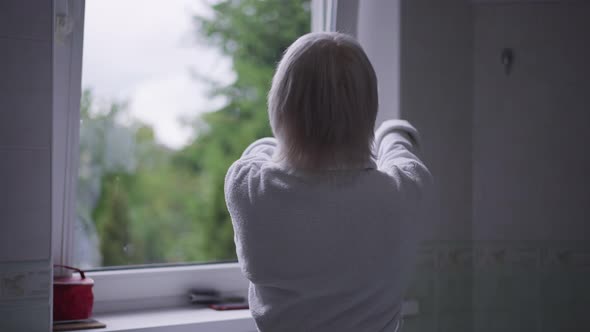 Back View Confident Caucasian Senior Woman with Grey Hair in White Bathrobe Stretching Looking Out