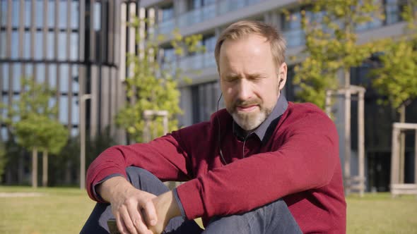 A Middleaged Handsome Caucasian Man Listens to Music with Earphones on a Smartphone As He Sits