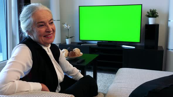 Old Caucasian Woman Watches Television in Living Room and Then Smiles To Camera