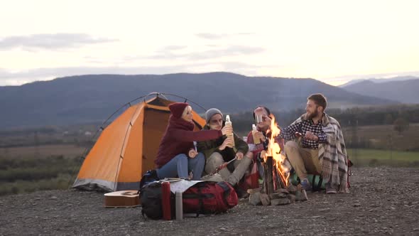 friends having rest near tourist tent with bonfire on which fried sausages in mountains