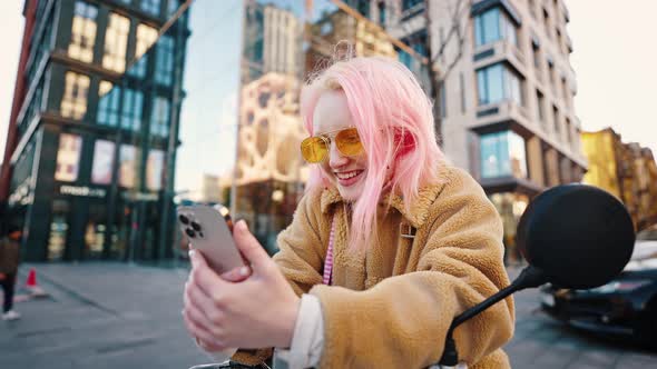 Young Positive Woman with Pink Hair Networking on Smartphone Sitting on Scooter Outdoors Tracking