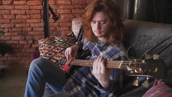 Portrait of Young Man Musician with Long Red Hair Playing Bass Guitar at Home Sitting on Sofa