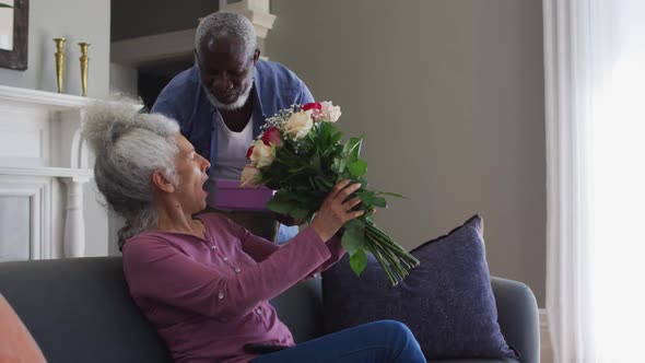 African american senior man giving gift box and flower bouquet to his wife at home