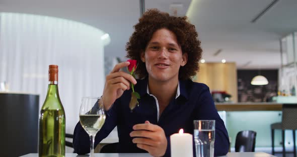 Mixed race man having romantic dinner at restaurant holding red rose and talking to camera