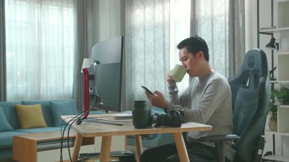 Asian Cameraman Using Mobile Phone And Drinking Coffee While Using Computer For Working At Home