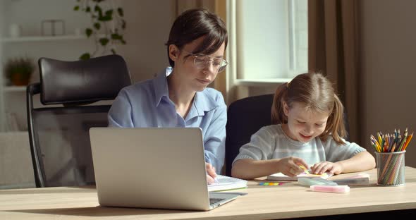 Young Woman Teacher Educator Nanny Wears Glasses, Sits with Little Girl at Table, Checks Homework