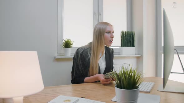 Woman Celebrating Success in Office. Elegant Blond Female Sitting at Workplace Holding in Hands