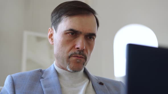 A Man with a Beard Sits in a Jacket on the Sofa Typing on a Laptop Keyboard