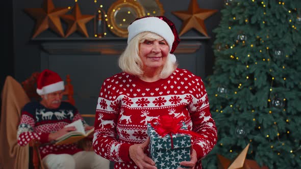 Mature Grandmother in Festive Sweater Presenting Christmas Gift Box Smiling Looking at Camera