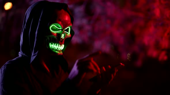 Portrait of a Person in a Glowing Mask of Death and in a Hood Against a Background of Branches