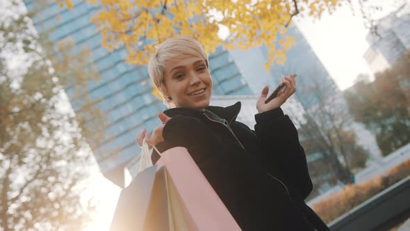 Young Woman Shopaholic Standing in the Park with Shopping Bags and Smartphone and Smiling