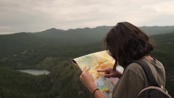 A young girl on a trip stands with a paper map against the background of mountains and a lake.