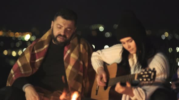 Group of people having fun sitting near bonfire outdoors at night playing guitar, singing songs 